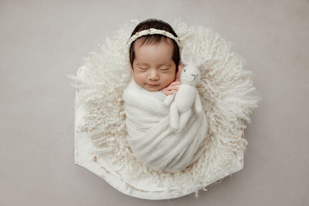 Newborn baby wrapped in a white blanket, lying on a fluffy white cushion, holding a small stuffed animal and wearing a headband—perfectly captured by a skilled newborn photographer.