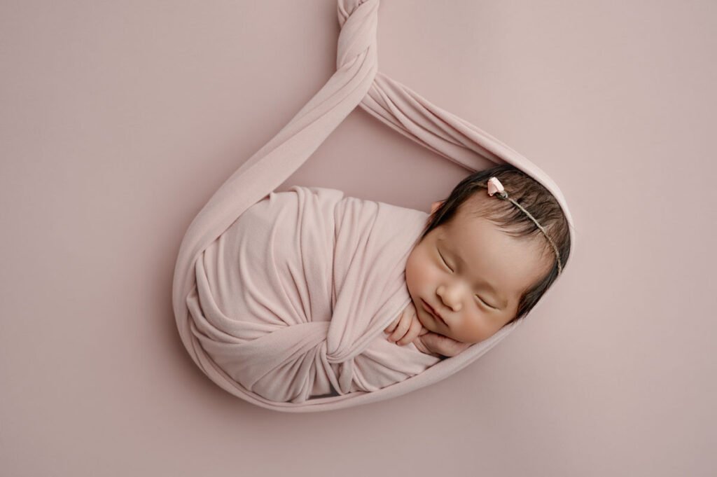 Newborn photographer captures a precious moment: a newborn baby wrapped in a soft pink blanket, sleeping peacefully on a matching pink background with a small pink hairclip.