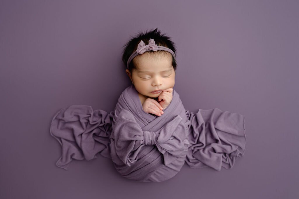 Newborn baby wrapped in a purple blanket with a bow, lying on a purple surface, wearing a matching headband, and sleeping with hands gently crossed under the chin—perfectly captured by a skilled newborn photographer.