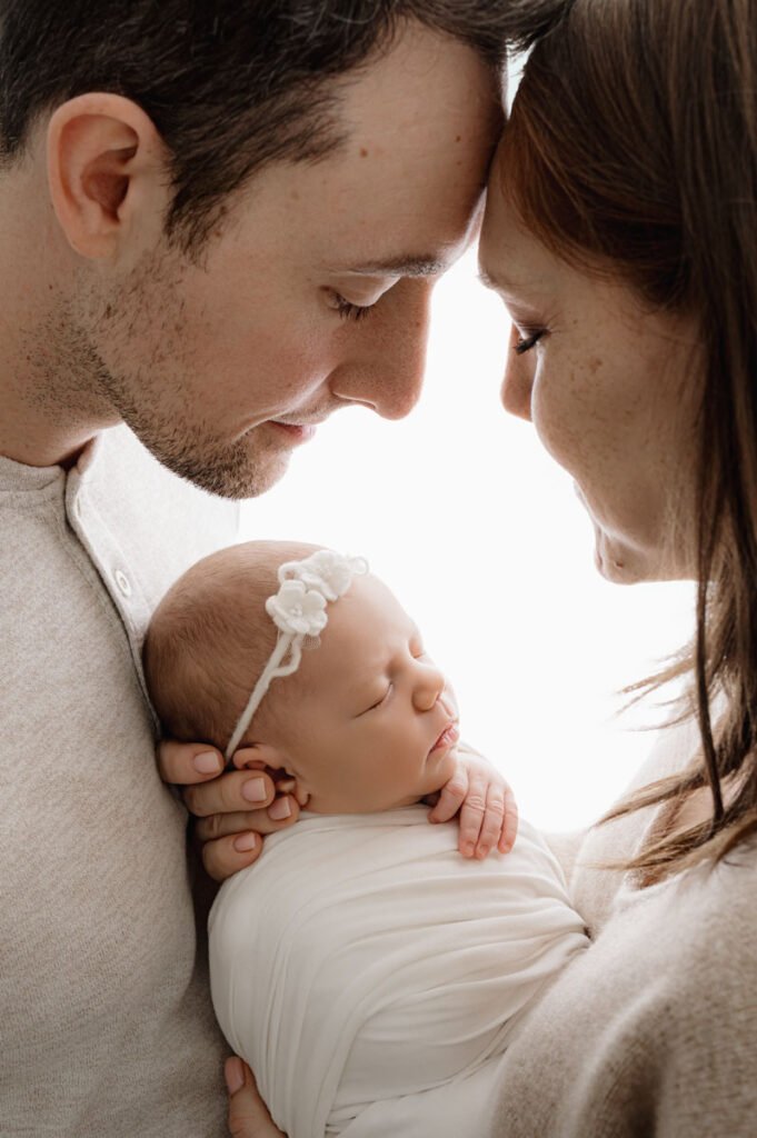 A man and woman gently touch foreheads while holding a swaddled newborn baby, who is wearing a white headband with flowers—a heartwarming moment beautifully captured by a skilled newborn photographer.