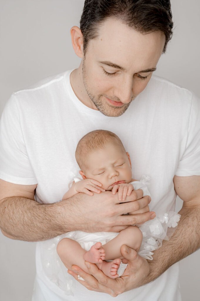 A newborn photographer captures a tender moment as a man in a white shirt gently cradles a sleeping newborn baby wrapped in white fabric, both facing the camera against a neutral background.