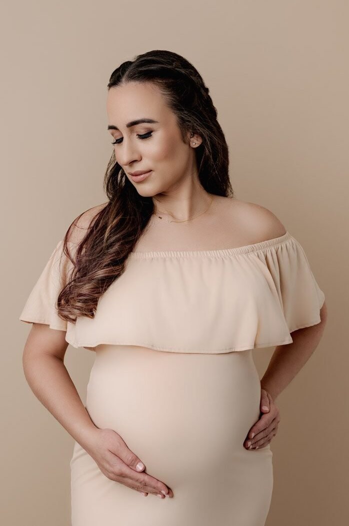 Pregnant woman in a soft beige off-shoulder dress cradling her belly, maternity portrait in North Vancouver