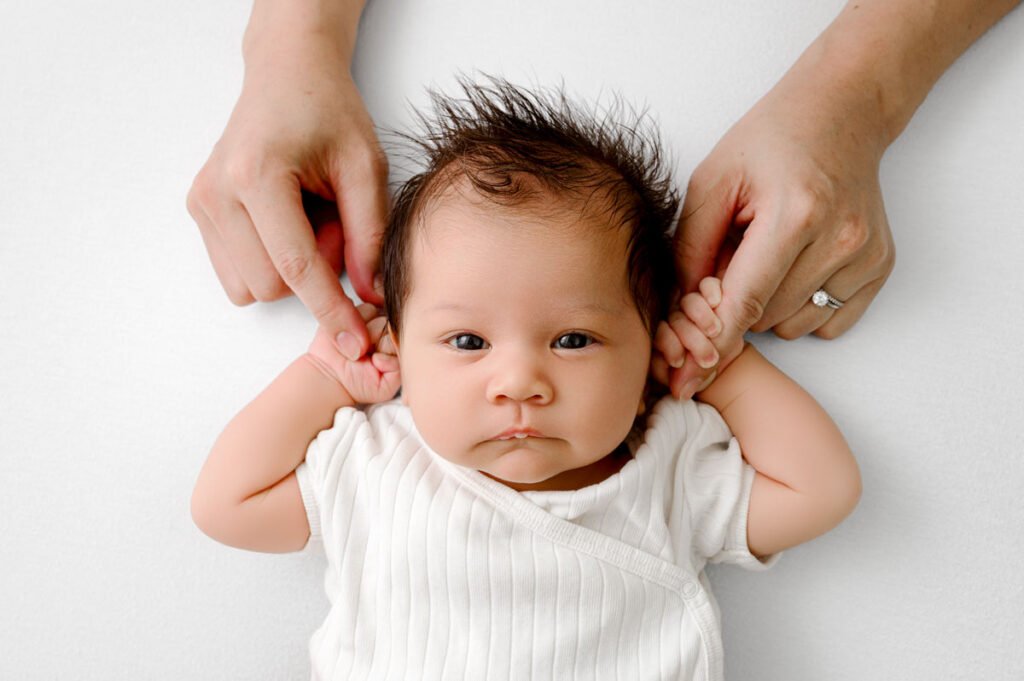 An adult’s hands gently hold a newborn baby’s hands up by their head.