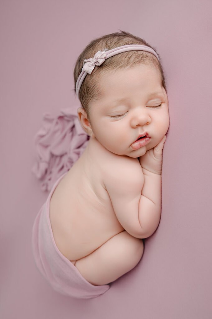 Newborn baby sleeping on a pink background, wearing a small bow headband and wrapped in a pink cloth.