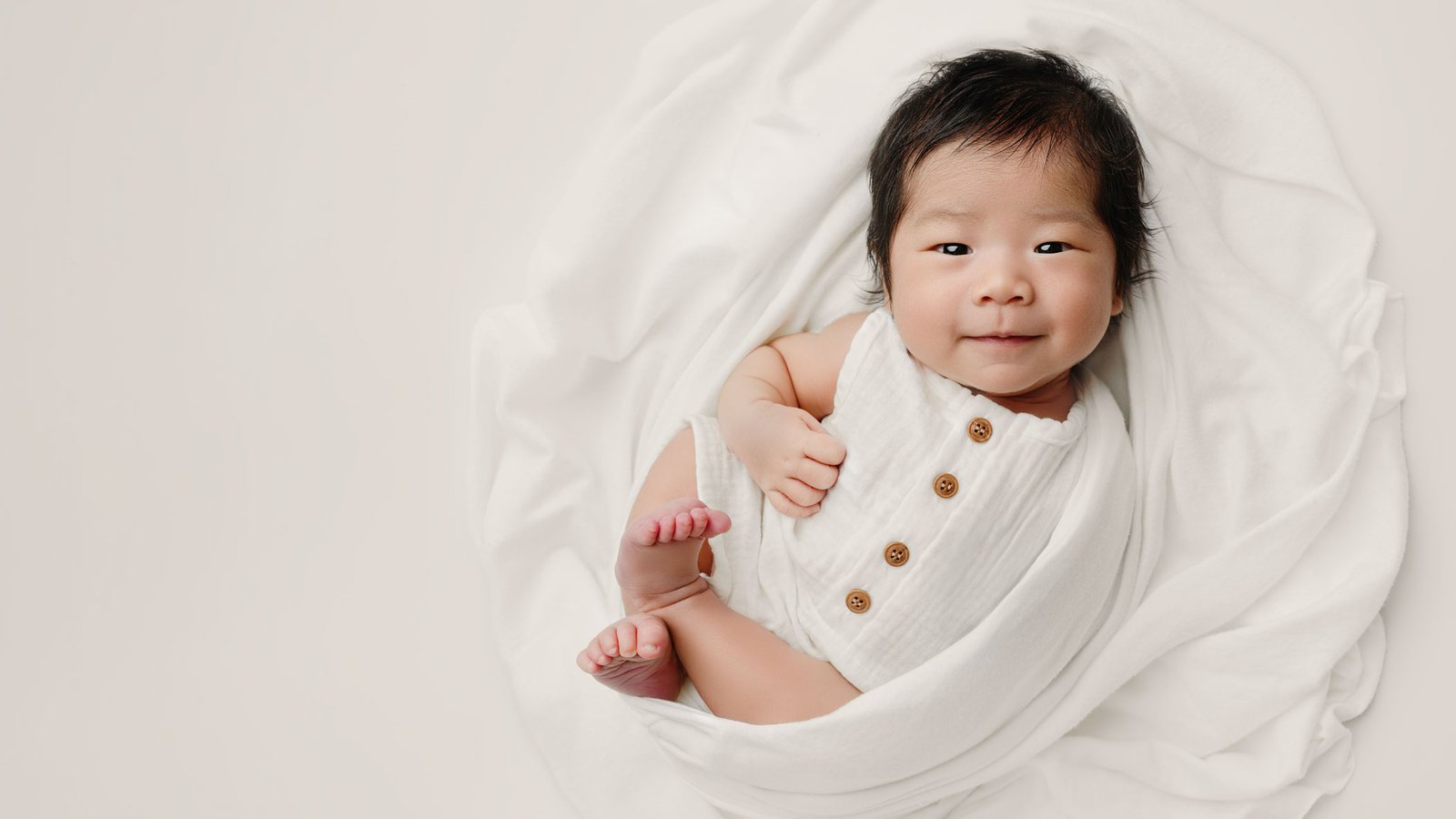 An awake older newborn baby in a white button-down onesie lying on a cream cloth.