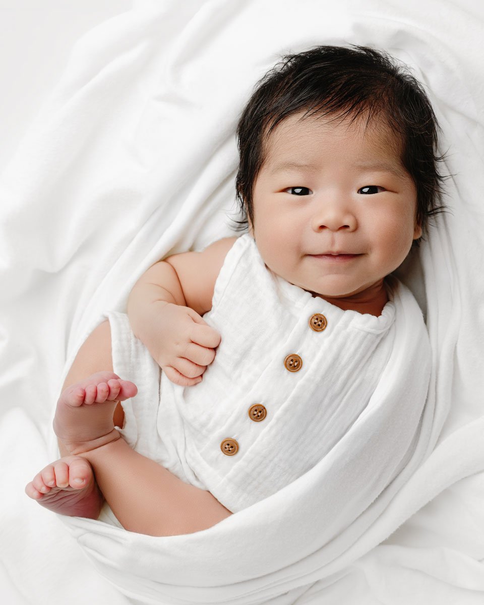 Smiling baby in a white button romper on a soft white blanket, North Vancouver newborn photography