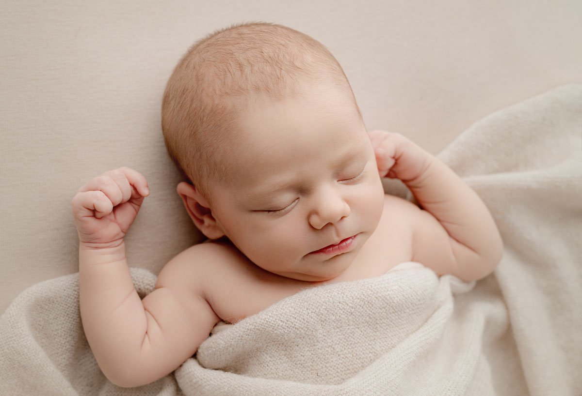 A newborn baby sleeping, wrapped in a soft blanket with arms raised.