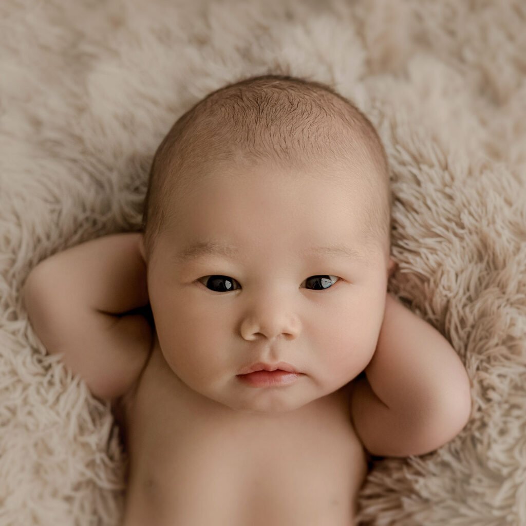 A newborn baby lying on a fuzzy surface with their hands behind their head.